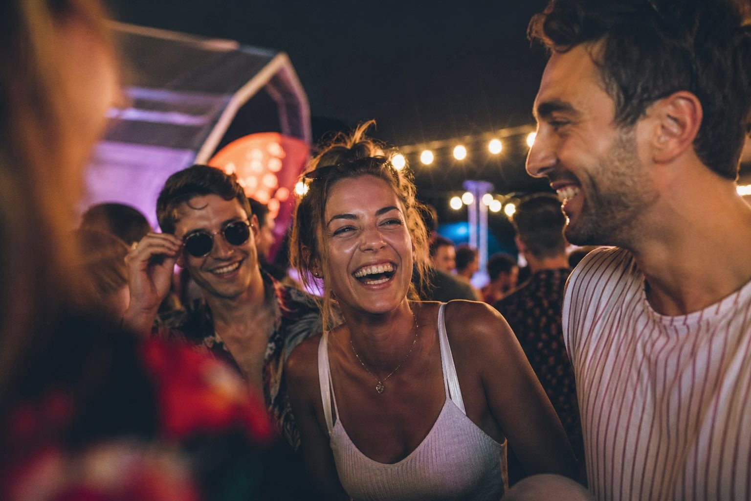 Friends laughing together at a summer evening event in the city, warm string lights overhead